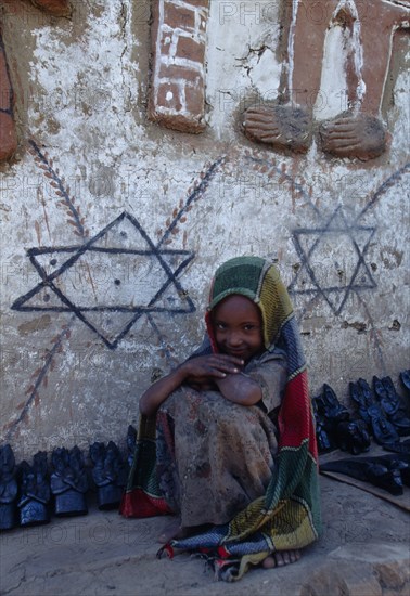 ETHIOPIA, Gonder Province, Felasher Village, Smiling young girl crouched beside typically decorated house and traditional pottery.