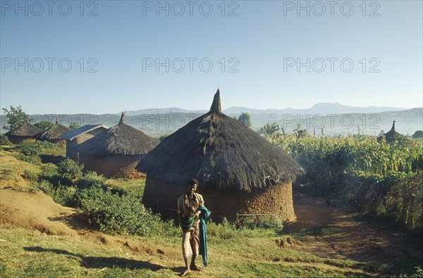 ETHIOPIA, Harerge Province, "Village near city of Harer.  Circular, thatched huts with villager holding a baby standing in the foreground."
