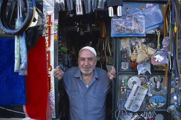 SYRIA, North, Halab, Male shopkeeper at the entrance to his shop with display of mixed goods hanging from the walls and ceiling.