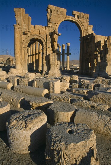 SYRIA, Central, Tadmur, Monumental arch. High central arch flanked by lower arch on each side with colonnaded street partly seen behind and masonry ruins in the foreground.