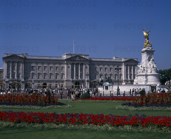 ENGLAND, London, Buckingham Palace and the Victoria Memorial with crowds of visitors outside.  Formal flowerbeds with red and blue flowers in the foreground.