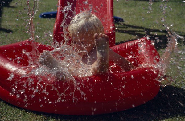 20016668 SPORT Water Paddling Pool Young boy splashing in a red inflatable paddling pool.