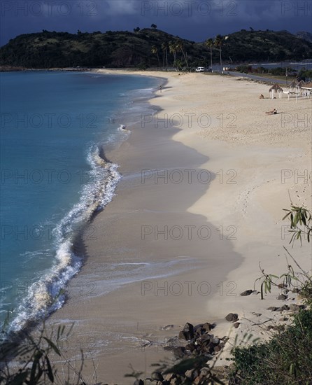 WEST INDIES, Antigua, Darkwood Bay, View along quiet sandy beach and bay with a few people sunbathing with road towards tree covered headland behind.
