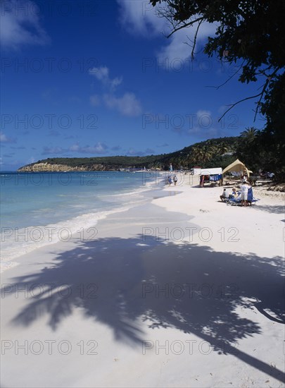 WEST INDIES, Antigua, Dickenson Bay, View along quiet sandy beach towards sunbathers and beach stall with headland beyond.  Shadow from palm tree in the foreground.