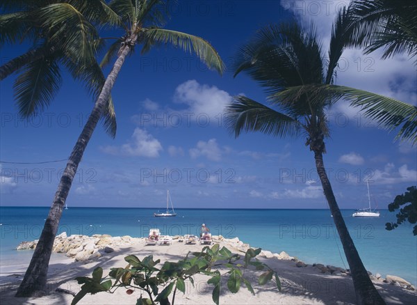 WEST INDIES, Antigua, Dickenson Bay, Sandy beach and rocks with people sunbathing on loungers and man fishing.  Aquamarine water and yachts framed by palms in the foreground.