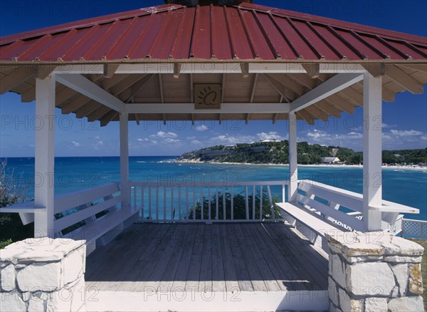 WEST INDIES, Antigua, Long Bay, Wooden decking with red painted roof and white seating framing view out over the bay and beach.