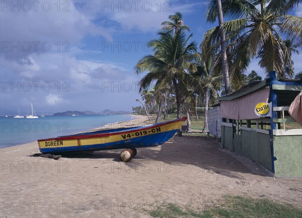 WEST INDIES, Nevis, Pinneys Beach, Empty sandy beach fringed by palm trees with painted wooden boat pulled up out of the water beside empty hut advertising Carib beer.