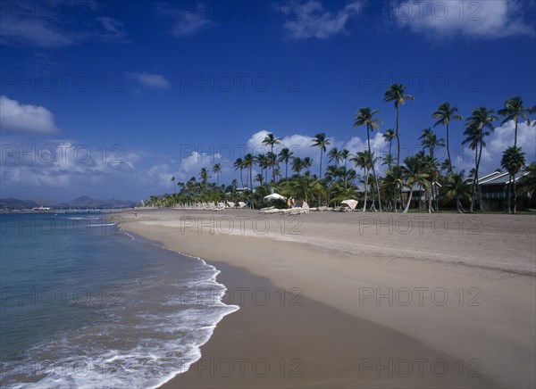WEST INDIES, Nevis, Pinneys Beach, Four Seasons Resort.  Quiet sandy beach at the waters edge.  Line of empty sun loungers with apartment buildings partly seen through palm trees behind.