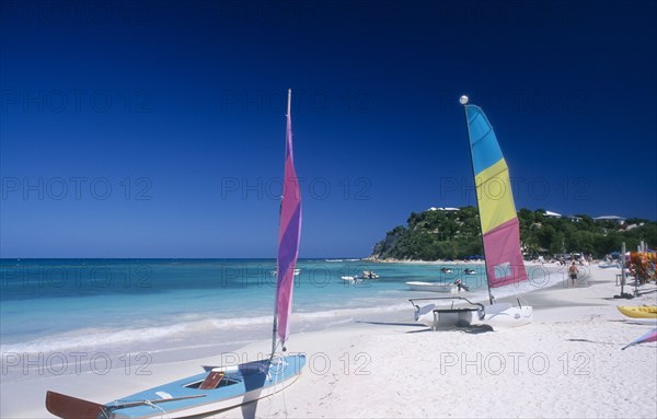 WEST INDIES, Antigua, Long Bay, "Semi circular bay beside quiet beach, boats with colourful sails pulled up onto the sand and motor boats and jet skis moored in the water.  Tree covered headland behind. "