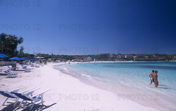WEST INDIES, Antigua, Long Bay, Sandy beach beside bay with blue and white deckchairs and couple standing in shallow water in the foreground.  Coastline with trees and buildings behind.