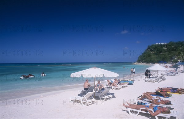 WEST INDIES, Antigua, Long Bay, Beach overlooking semicircular bay with sunbathers on white sun loungers.  Two jet skis in the water and tree covered headland behind.