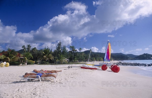 WEST INDIES, Antigua, Jolly Beach, Sandy beach fringed with palm trees with sailing boat and beach-bicycle on the shoreline and three sunbathers on white loungers in the foreground.