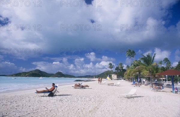 WEST INDIES, Antigua, Jolly Beach, View along sandy beach lined with palm trees and sun umbrellas towards hilly coastline. People sunbathing on loungers in the foreground.