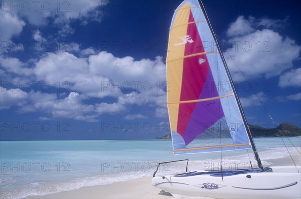 WEST INDIES, Antigua, Jolly Beach, Empty sandy beach with hobiecat with brightly coloured sail in the foreground.  Flat calm aquamarine sea and cloudscape behind.