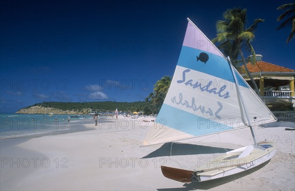 WEST INDIES, Antigua, Dickenson Bay, View along sandy beach lined with palms with boat in the foreground advertising Sandals hotel on its hull and sail.  Building rooftop part seen behind.