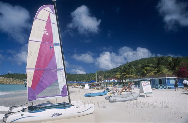 WEST INDIES, Antigua, Dickenson Bay, "Sandy beach with people on deck chairs beside blue painted building, A-board advertising Tonys watersports.  Hobiecat with bright sail in front."