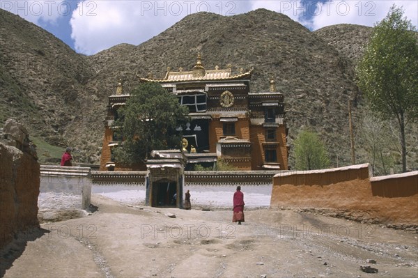 CHINA, Gansu, Xiahe, "Labrang Monastery, elaborately designed building with a golden roof, partially obscured by a tree, and monks walking in the foreground."
