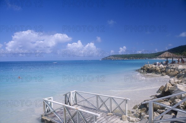 WEST INDIES, Antigua, Dickenson Bay, Raised area of sand fortified by rocks with people sitting on loungers under thatched umbrella with man swimming and wooden jetty in the foreground.