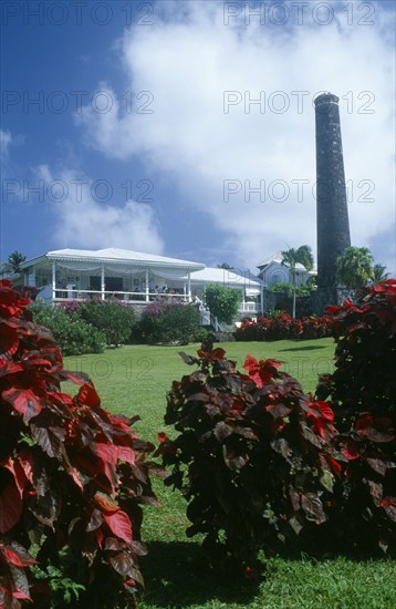 WEST INDIES, St Kitts, Rawlins Plantation Inn, Exterior with people on white painted balcony and old sugar factory tower and wall on the right.  Red and purple variegated plants in the foreground.