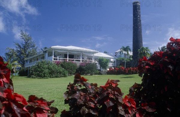 WEST INDIES, St Kitts, Rawlins Plantation Inn, Exterior with people on white painted balcony and old sugar factory tower and wall on the right.  Red and purple variegated plants in the foreground.