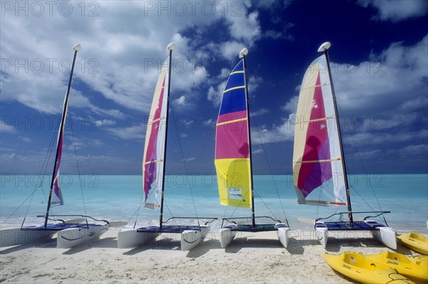 WEST INDIES, Antigua, Jolly Beach, Line of hobiecats with brightly coloured sails on sandy beach.  Sea behind and blue sky with windswept clouds.