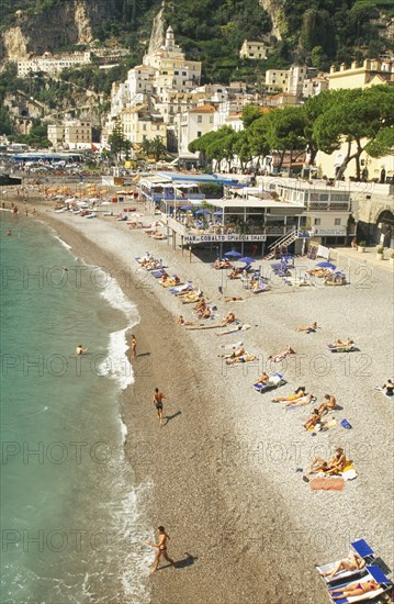 ITALY, Campania, Amalfi Coast, Amalfi.  View along shingle beach with sunbathers and cafes towards town buildings on hillside.