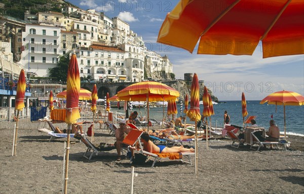 ITALY, Campania, Amalfi Coast, Amalfi.  Beach with sunbathers on loungers under yellow and orange striped beach umbrellas.  Town buildings on the hillside behind.