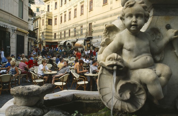 ITALY, Campania, Amalfi Coast, Amalfi.  Street scene with people seated at outdoor tables of busy cafe.  Detail of fountain stonework in the foreground.