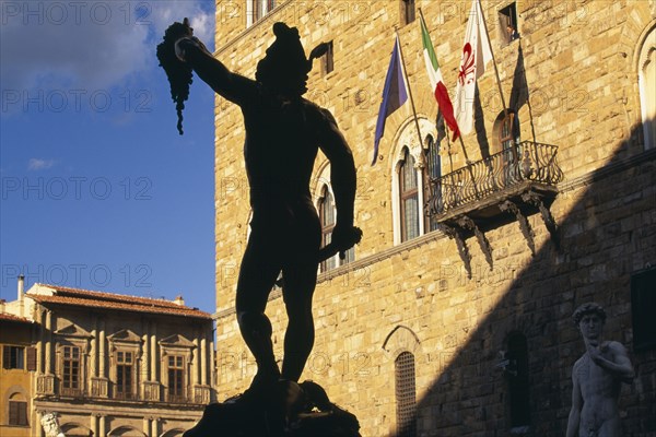 ITALY, Tuscany, Florence, Piazza della Signoria.  Statue of Perseus holding the head of Medusa by Cellini silhouetted against Palazzo Vecchio in sunlight.