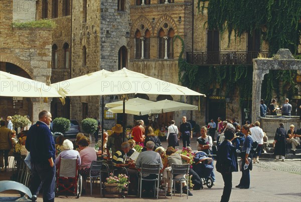 ITALY, Tuscany, San Gimignano, Street scene with people sitting at outside tables of busy cafe with sun umbrellas.
