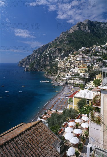ITALY, Campania, Positano, View over balcony with tables and chairs under white sun umbrellas towards beach and village houses built on steep hillside beyond