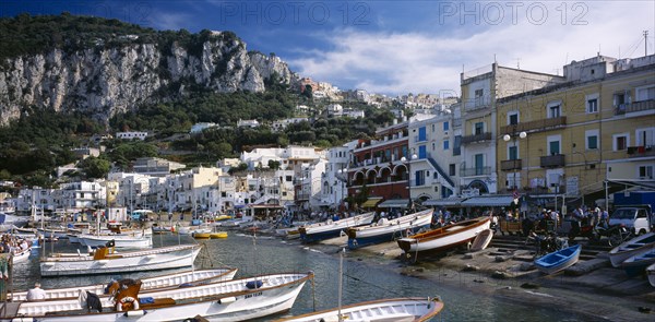 ITALY, Campania, Capri, Marina Grande. View over moored boats in harbour lined with colourful houses and busy cafes and restaurants. Rocky hillside behind.