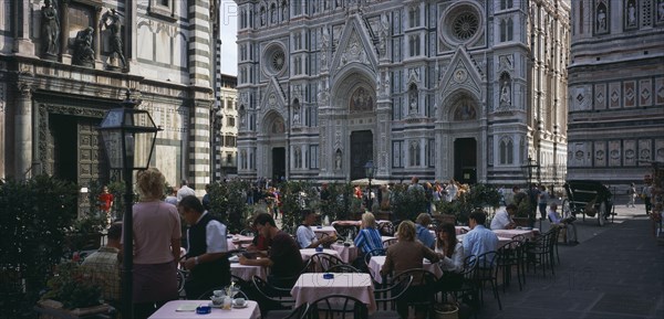 ITALY, Tuscany, Florence, Cafe in front of the Neo-Gothic marble facade of the Duomo with people sitting at outside tables having coffee and reading guide books.