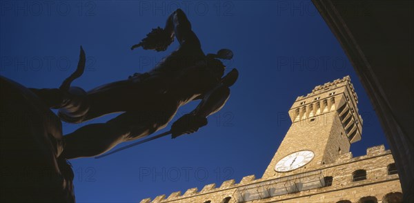 ITALY, Tuscany, Florence, Piazza della Signoria.  Angled view of statue of Perseus with the head of Medusa by Cellini in silhouette with Palazzo Vecchio in sunlight behind.