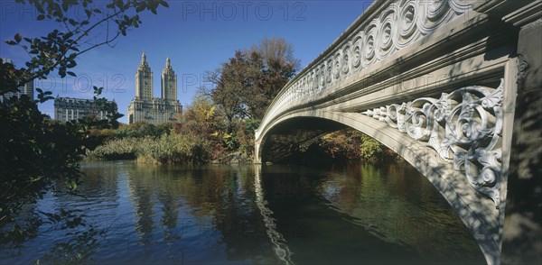 USA, New York , Manhattan, "Central Park, Bow Bridge.  View along side of cast iron bridge across lake lined by trees and overlooked by city buildings and the San Remo towers ."