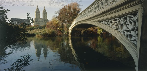 USA, New York , Manhattan, "Central Park, Bow Bridge.  View along side of cast iron bridge across lake lined by trees and overlooked by city buildings and the San Remo towers."