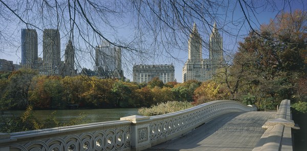 USA, New York State, New York, "Central Park, Bow Bridge.  View over cast iron bridge across lake lined by trees in autumn colours, overlooked by city buildings and the San Remo towers."