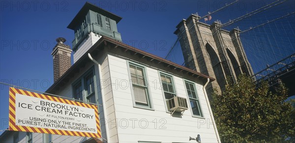 USA, New York, Manhattan, Part view of Brooklyn Bridge seen above trees with Brooklyn Ice Cream Factory building and sign in the foreground.