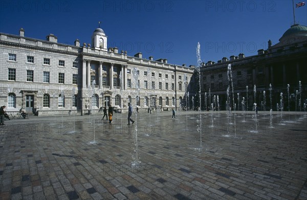 ENGLAND, London, Somerset House and courtyard with rows of fountains spouting high from the ground with people walking between them. Flag flying at half mast in the distance.
