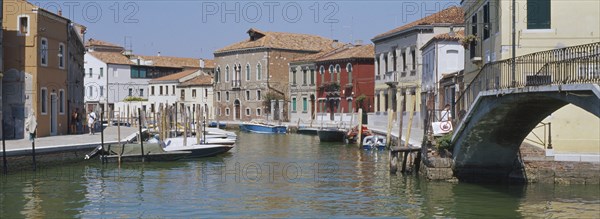ITALY, Veneto, Venice, Murano Island.  View along canal lined by traditional houses with boats moored to posts at each side.