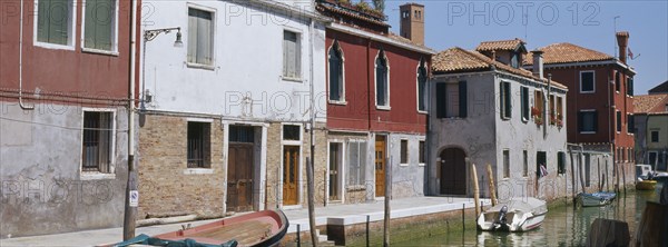 ITALY, Veneto, Venice, Murano Island.  View along canalside houses with boats moored to posts in front.
