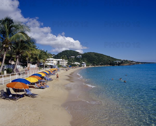 WEST INDIES, US Virgin Islands, St Thomas, Frenchman Bay.  View along sandy beach with row of sun loungers and brightly coloured beach umbrellas. People sunbathing and swimming.