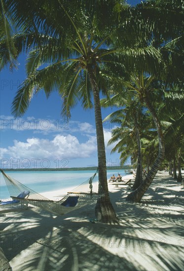 PACIFIC ISLANDS, Cook Islands, Aitutaki, Akitua.  Sandy beach with people sunbathing on wooden sun loungers and hammock slung between palm trees in the foreground.