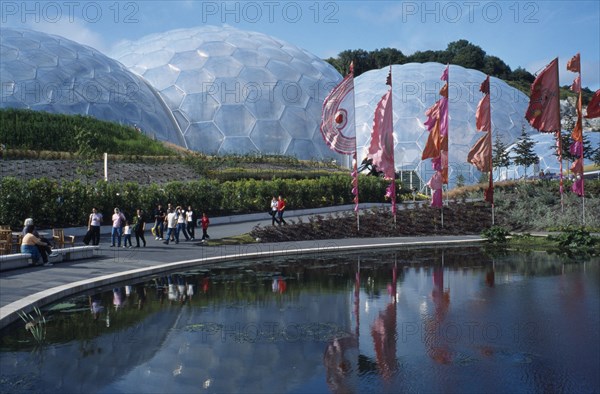 ENGLAND, Cornwall, St. Austell, Eden Project.  General view over the Humid tropics Biome exterior with line of coloured flags and visitors walking beside lake.