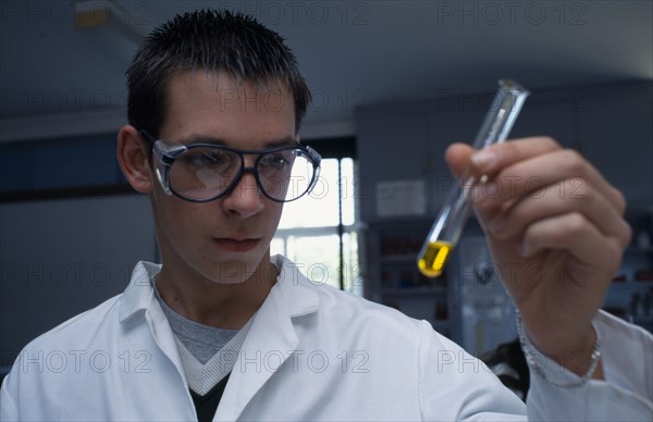EDUCATION, Science Class, Male student wearing protective glasses and lab coat holding a test tube containing yellow liquid at an angle.