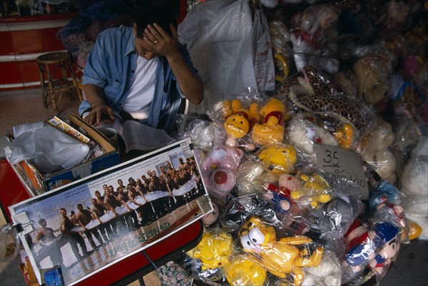 THAILAND, North, Chiang Mai, Chinatown. Shop with plastic wrapped toys for sale and picture commemorating the capture of a huge fish by the American Army in 1973.  Male vendor reading behind.