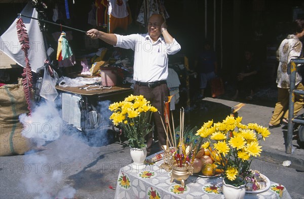 THAILAND, North, Chiang Mai, "Chinese New Year.  Man at roadside stall holding his ears as he sets off chain of fire crackers.  Table with flowers, incense and food offerings beside him."