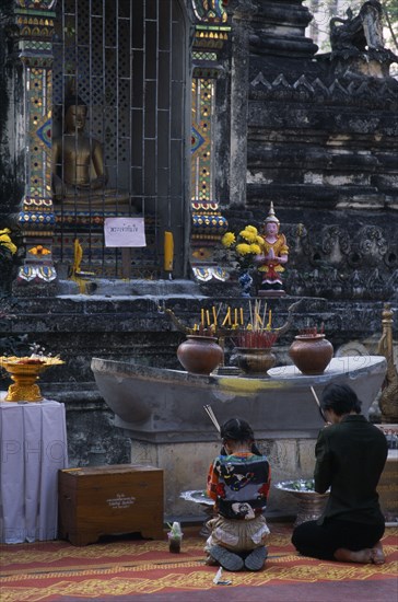THAILAND, North, Chiang Mai, Wat Chettawan on Tha Phae Road.  Young woman and girl kneeling with incense in front of stone niche with seated Buddha and altar with burning incense.