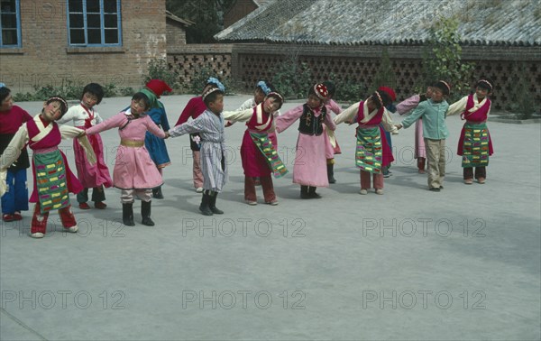 CHINA, Hebei Province, Chengde, Line of children at kindergarten with linked arms and heads inclined sideways wearing traditional dress.