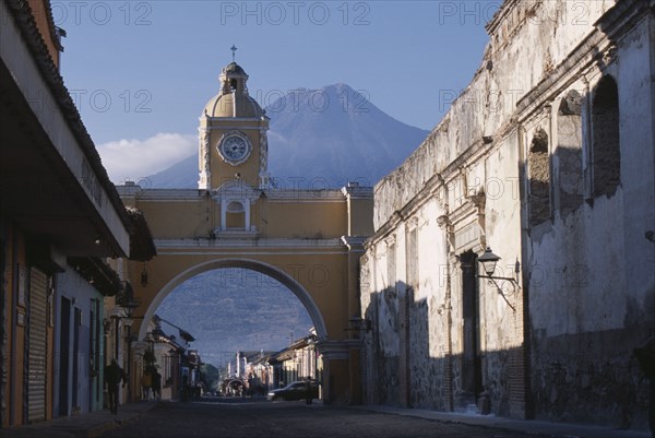GUATEMALA, Antigua, "Arco de Santa Catarina. Yellow painted archway with clock tower and people walking underneath, with Volcan Agua beyond."